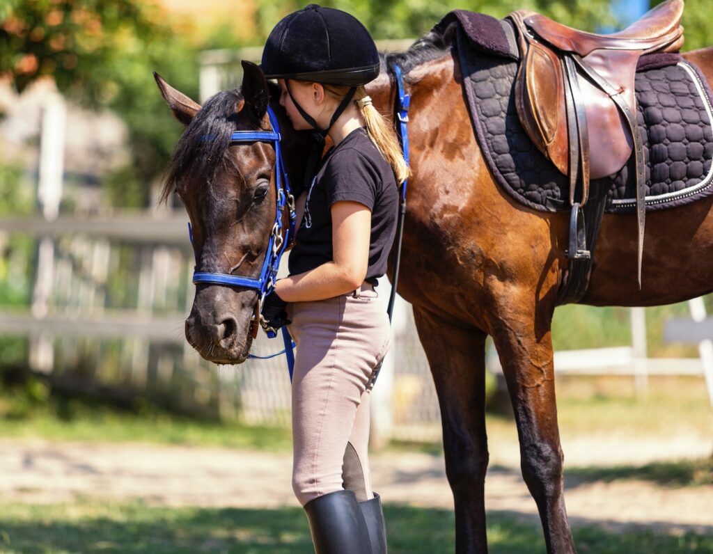 Beautiful girl in riding gear holds her horse by the reins. She stroking him after class at riding school.