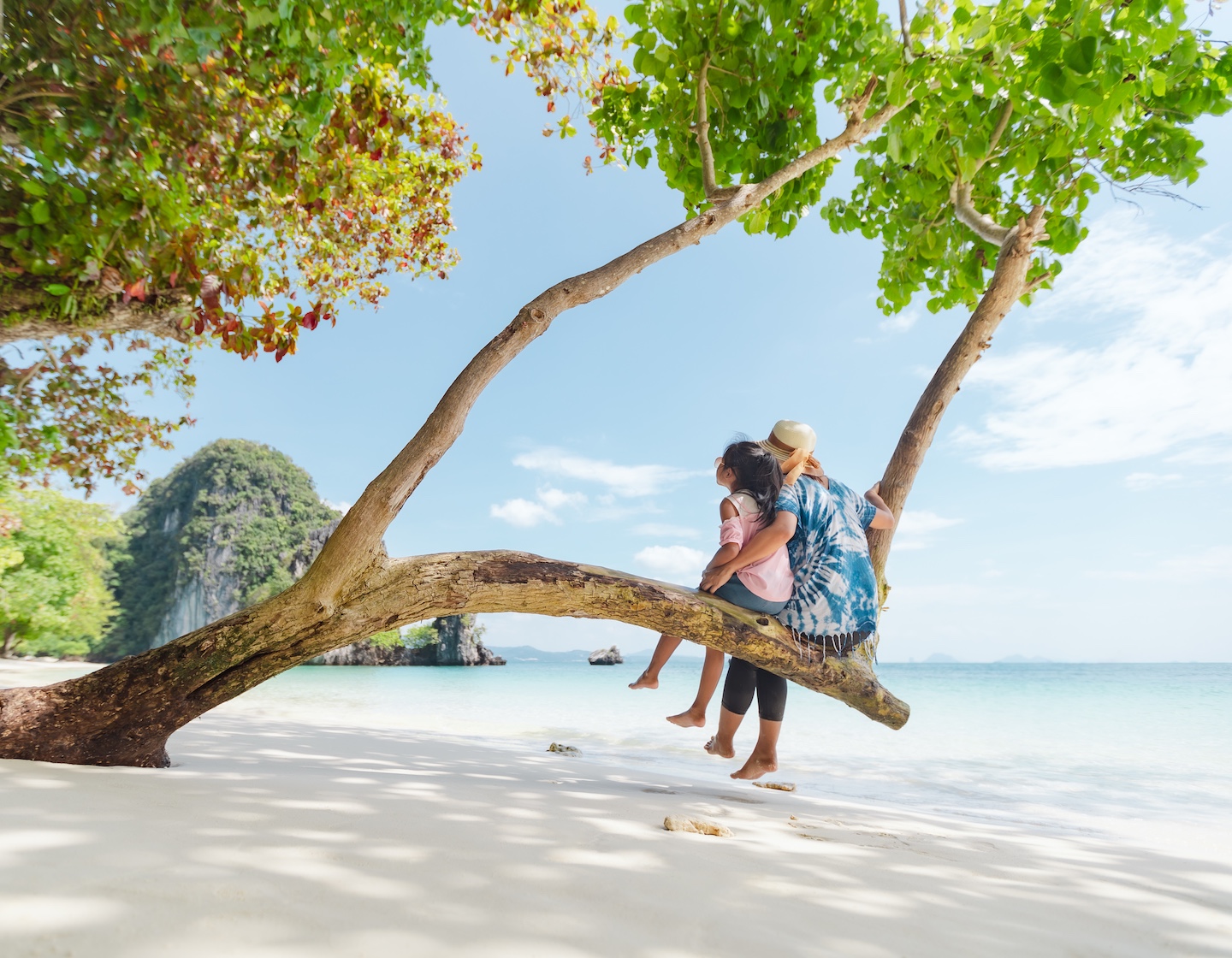 Asian mother and child girl sitting on tree and enjoying with beautiful sea nature together in their vacation. Summer holidays and family travel concept. Travelling in Thailand.