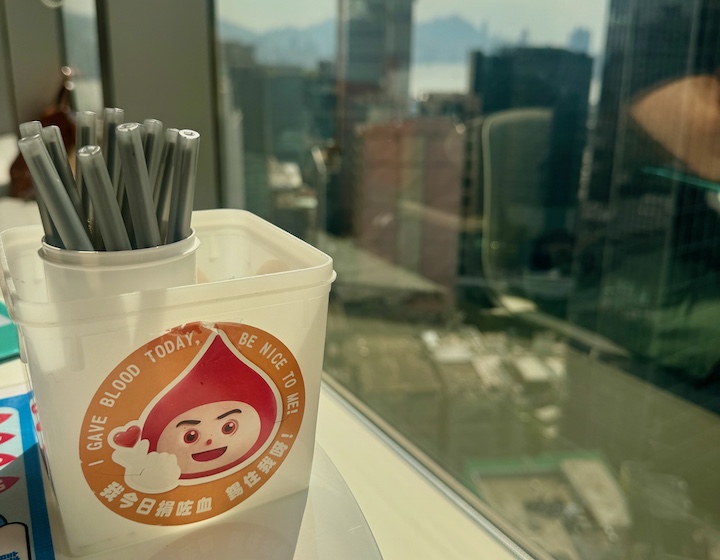 a container of pens with a blood donation sticker on it, with the window overlooking Kwun Tong 
